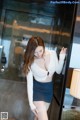 A woman in a white top and blue skirt leaning against a glass door.